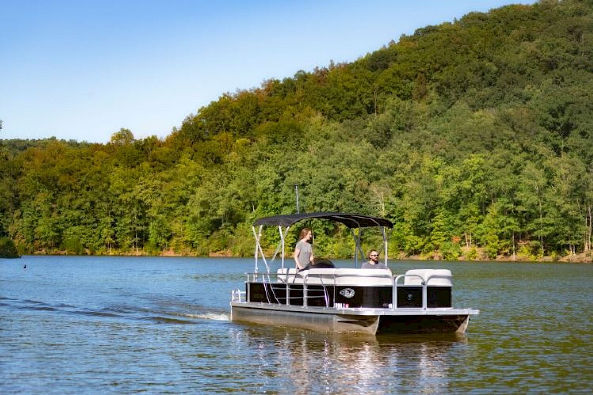 A pontoon boat with two people is cruising on a lake surrounded by lush green trees and hills under a clear blue sky.