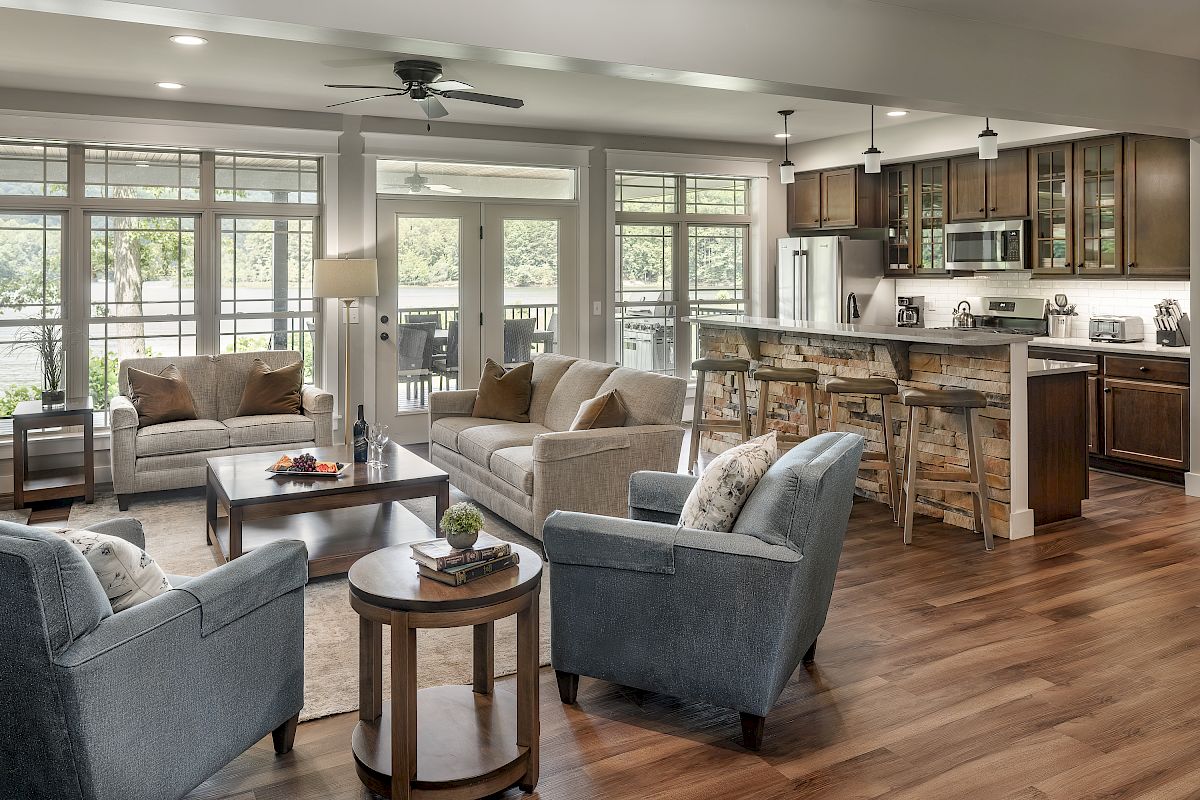 A modern living room and kitchen area with grey and beige furniture, wooden flooring, a ceiling fan, and large windows letting in ample light.