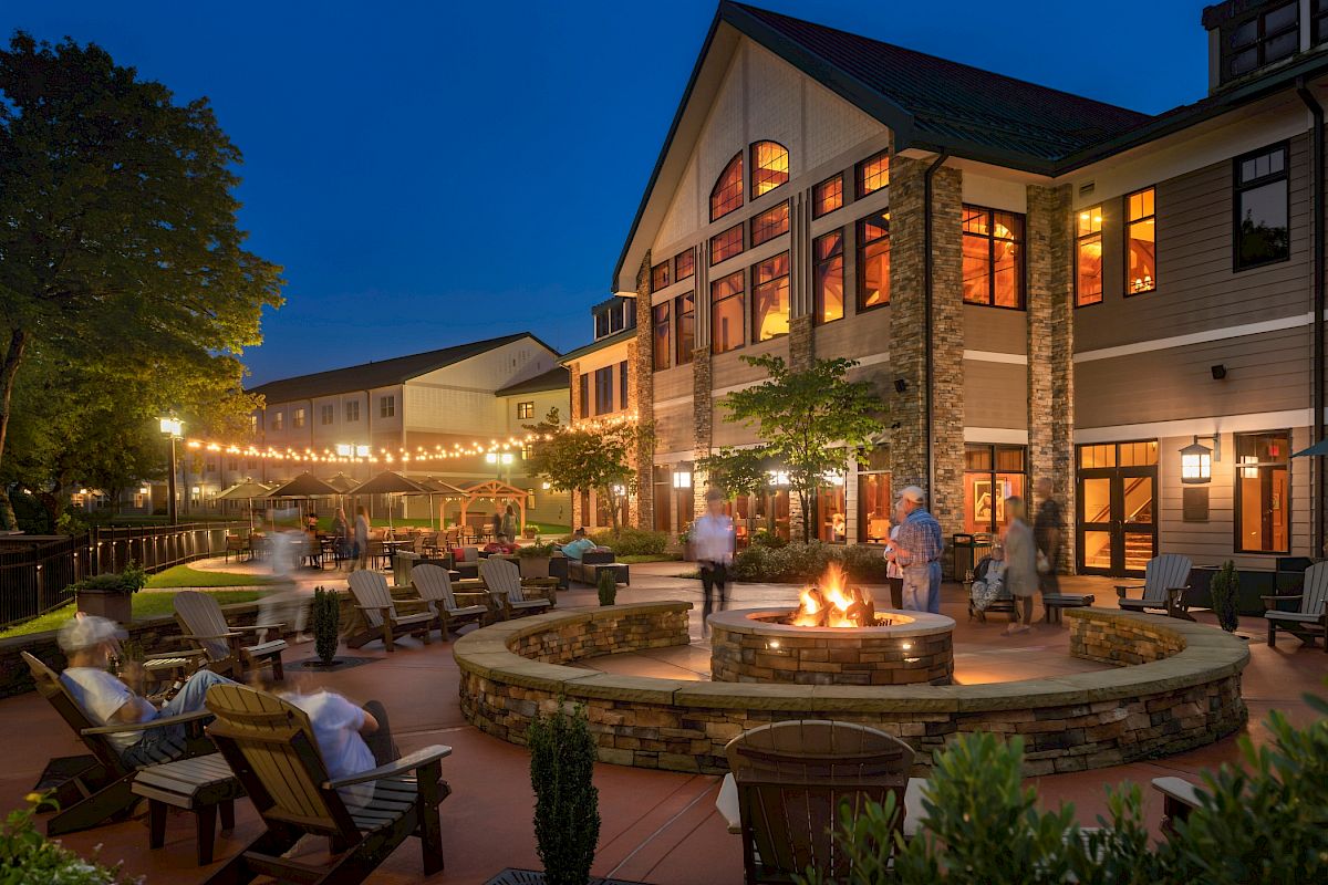 An outdoor evening scene shows people relaxing around a fire pit at a resort-like facility with string lights, trees, and a spacious building.